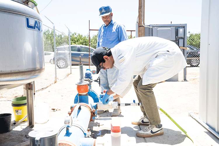 On a bright sunny day, a person wearing a white lab coat and a black baseball cap leans over to collect a water sample from an outdoor well. A second person, wearing a blue denim shirt, stands behind them.