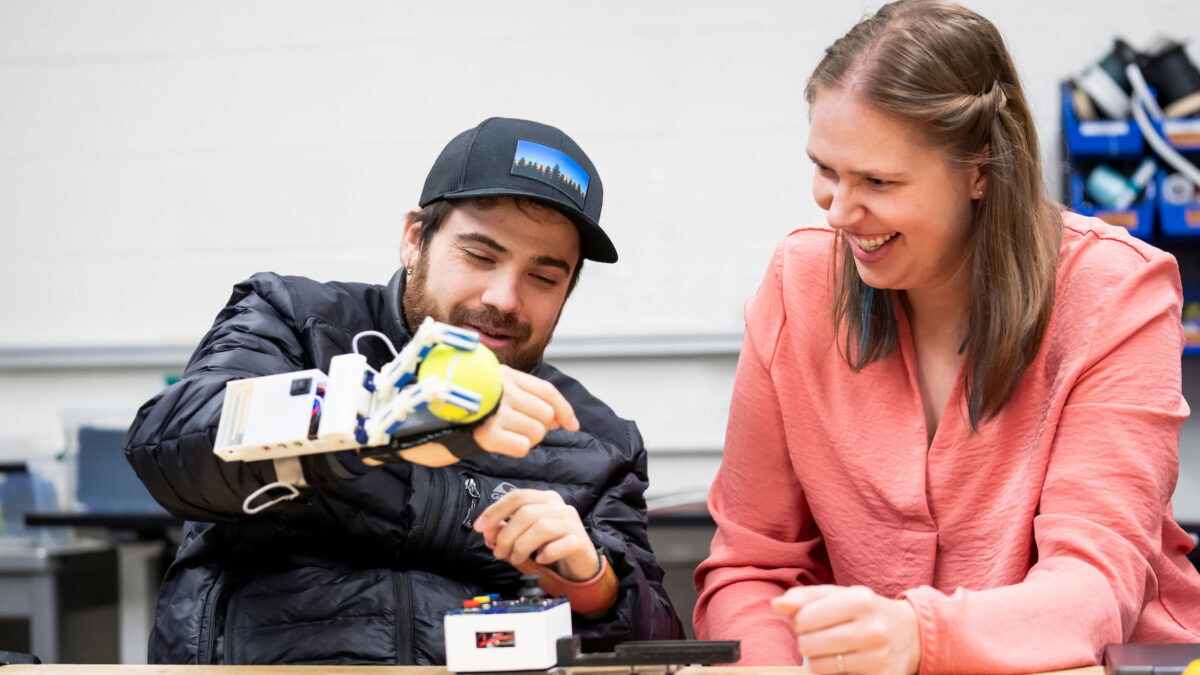 A researcher demonstrates a robotic device attached to the back of their hand as it grasps a tennis ball.